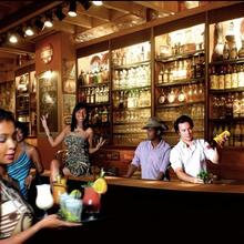 Interieur Hotsy Totsy. Twee barmannen staan achter bruine toog cocktails te shaken, en enkele dames staan voor de toog te dansen. Centraal in de foto: een ober met verschillende cocktails op een plateau.