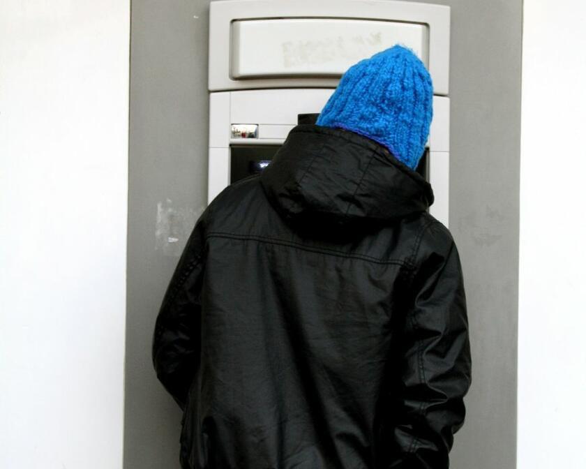 Un homme portant un manteau noir, un chapeau bleu et un jean prend un portefeuille dans sa poche arrière. Il se tient devant un distributeur de billets. En lettres bleues sur fond jaune, on lit "cash" au-dessus de la machine.