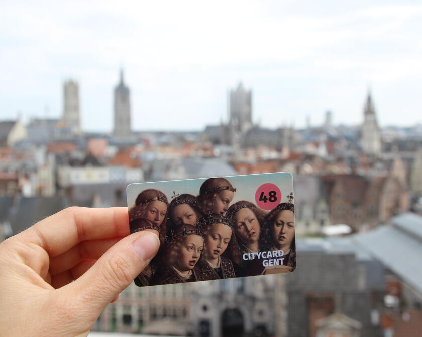 Una mano sujeta la City Card Gent (48h) con una vista panorámica de Gante de fondo