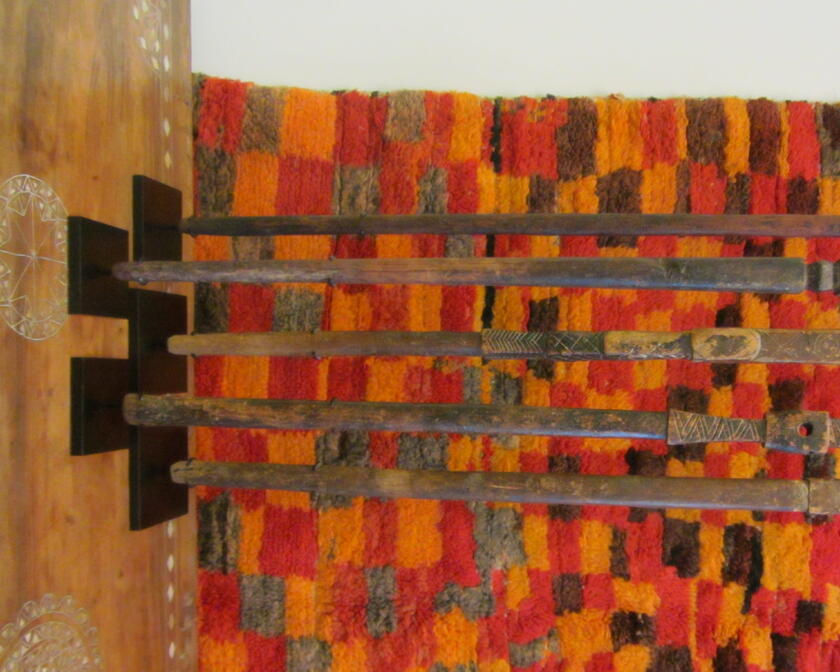 5 houten kunstwerken met een gat in voor een rechtstaand tapijt met oranje, bruine en rode rechthoekjes