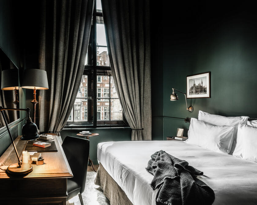 Tweepersoonskamer met kingsize bed, diepgroene muren en grijze gordijnen. Door het raam zie je een iconische gevel in de stad. 