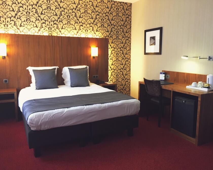 Tweepersoonskamer met houten bedeinde, rood tapijt op de grond en floraal behangpapier achter het bedeinde.