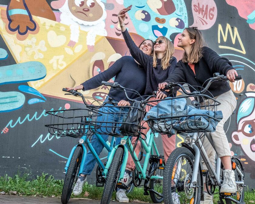 3 vrouwen die een selfie nemen op de fiets voor een graffitimuur