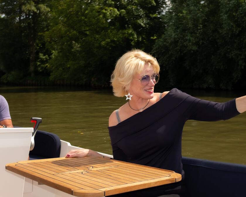 Pascale Platel met zonnebril die in een boot zit en naar iemand zwaait, de bootbestuurder
