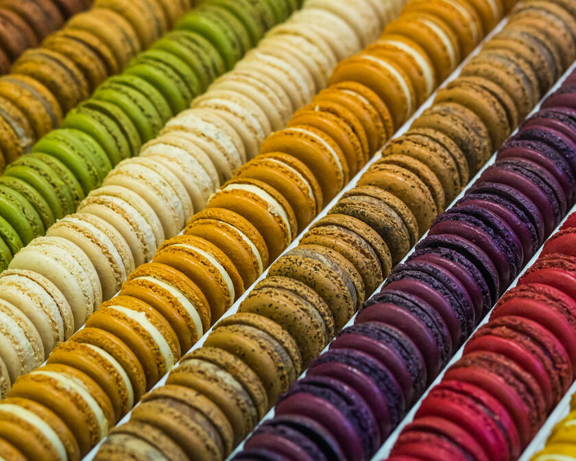 Verschillende rijen gekleurde macarons. Van links naar rechts: Bruin, groen, wit, oker, bruin met zwarte spikkels, paars en rood. 