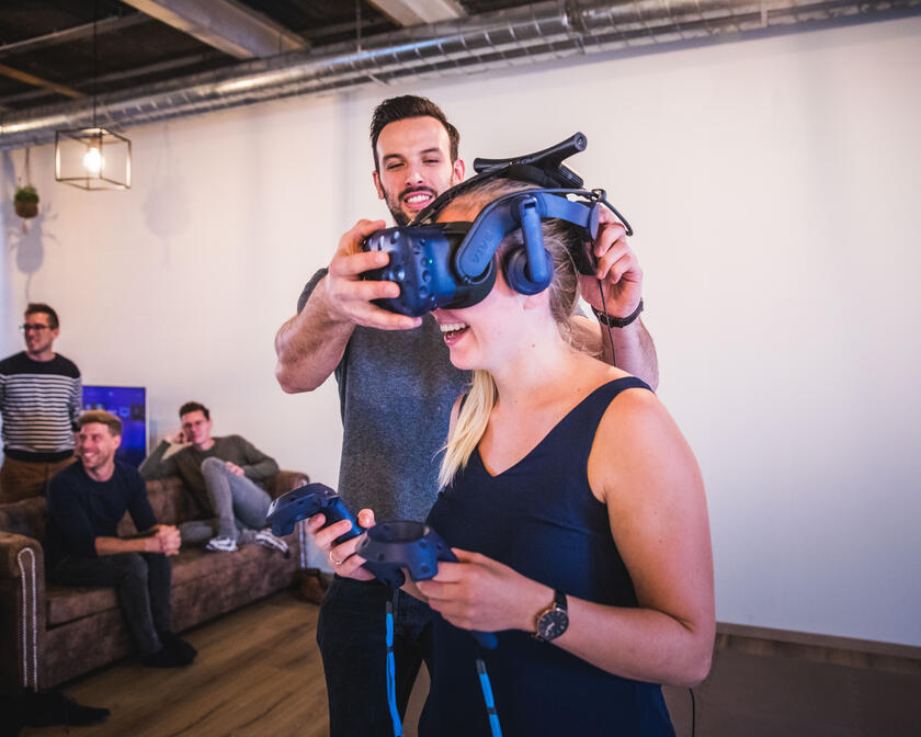 vrouw die een vr-bril uittest, man die hem op haar hoofd zet, mensen in een zetel die toekijken