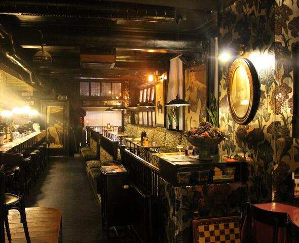 Lange ruimte met links de bar met krukken en rechts houten tafels met muurbanken en stoelen. Antiek behangpapier met grote bloemen.