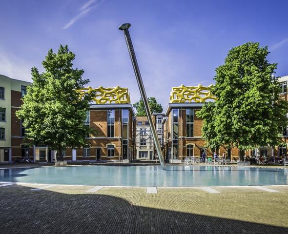La cour du complexe Zebrastraat dispose d'une terrasse où de nombreuses personnes sont assises. Des arbres couverts de feuilles s'élèvent à gauche et à droite. À l'avant se trouve un bassin dans lequel un clou géant a été enfoncé. Il s'agit de l'œuvre « Nagel » de Marnix Verstraeten. Sur le toit du complexe se trouvent deux structures à ossature jaune identiques. Il s'agit de l'œuvre WARSUBEC de Nick Ervinck.