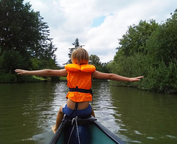 Kind mit Schwimmweste sitzt an der Spitze eines Kanus auf dem Fluss, umgeben von Grün.