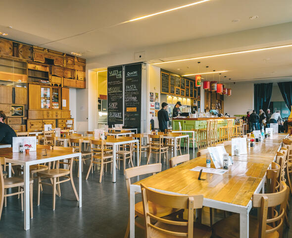 Interieur van het restaurant met houten tafels en stoelen, houten kastjes tegen de muur, menu op een groot krijtbord. Lampen gemaakt van oude, metalen dozen voor koffiebonen en lichtgroene toog. Klanten in de voor-en achtergrond, twee personeelsleden aan de toog.