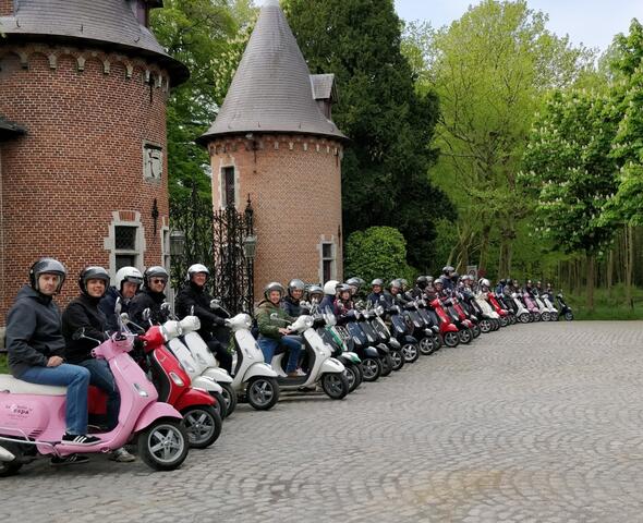 Een groep volwassenen op kleurrijke Vespa's die op een rij klaarstaan om te vertrekken. 