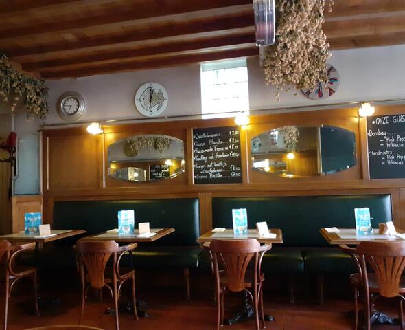 Interior del restaurante 't Klokhuys, donde se disponen cuatro mesas en fila, cada una con una silla de madera a un lado y un sofá verde al otro, frente a una pared de madera con espejos y dos pizarras en las que se indica la lista de precios de los postres y ginebras.