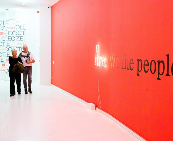 Two people walk past a red wall with 'Are we the people?' on it in the exhibition Public Matter(s) at Design Museum Gent.