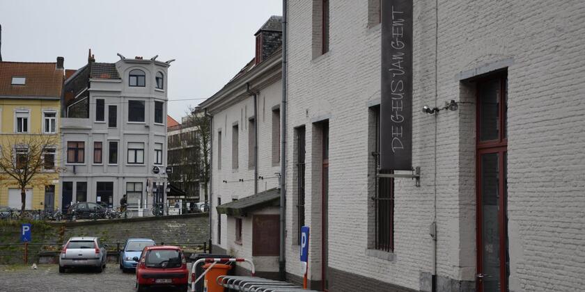 Voorgevel van de Geus van Gent. Witte, bakstenen voorgevel met fietsenstalling. Op de achtergrond zie je de huizen van de Muinkkaai.