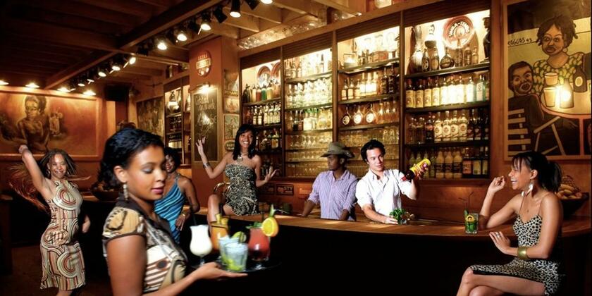 Interieur Hotsy Totsy. Twee barmannen staan achter bruine toog cocktails te shaken, en enkele dames staan voor de toog te dansen. Centraal in de foto: een ober met verschillende cocktails op een plateau.