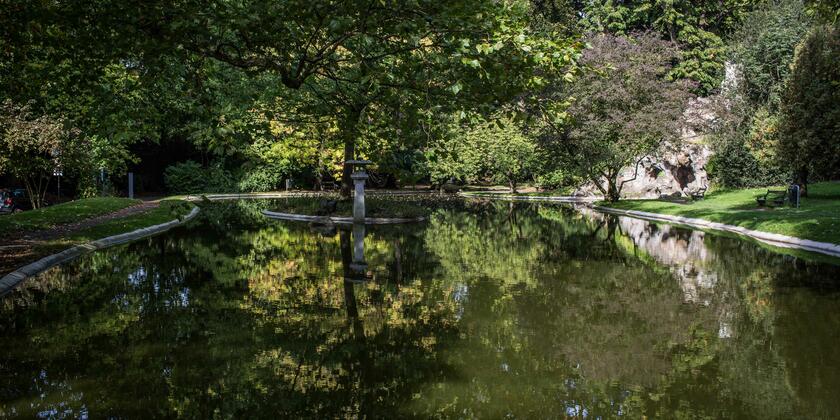 Photo idyllique depuis le Citadelpark. Au centre se trouve un étang, entouré de plusieurs arbres verts qui se reflètent dans l'eau. 