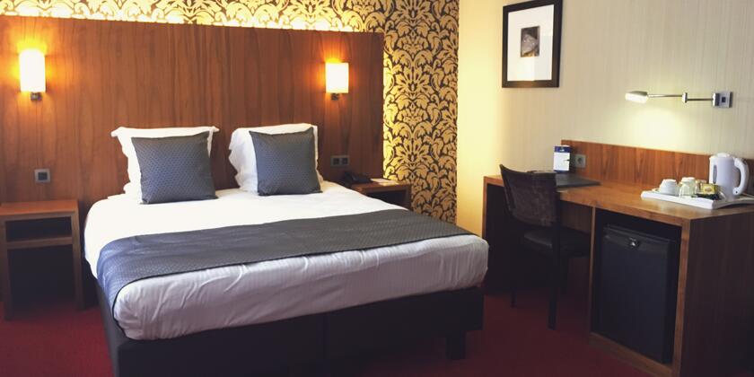 Tweepersoonskamer met houten bedeinde, rood tapijt op de grond en floraal behangpapier achter het bedeinde.