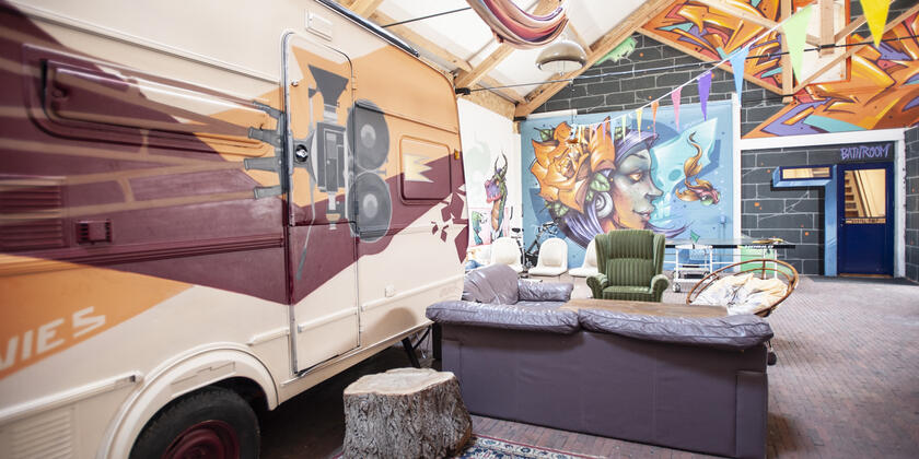 Interieur van Treck hostel Gent: een drukbeschilderde caravan staat binnen met salon ernaast. Op de achtergrond zie je de graffiti op de muren. 