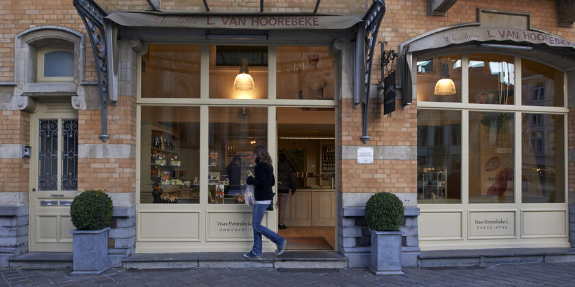 De gevel van de winkel, rode bakstenen, crèmekleurig houtwerk. Beige luifels (gesloten) met "L. Van Hoorebeke". Twee groene struiken in grijze, stenen potten. Een vrouw in een zwarte jas en blauwe jeans loopt langs de etalage.