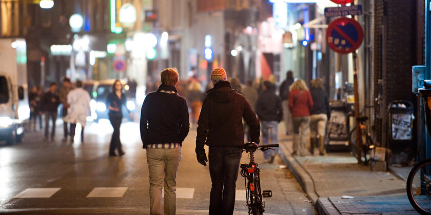 Twee jongeren (één met de fiets aan de hand) stappen weg van de fotograaf, richting een drukke straat met cafés en veel mensen; 's nachts.