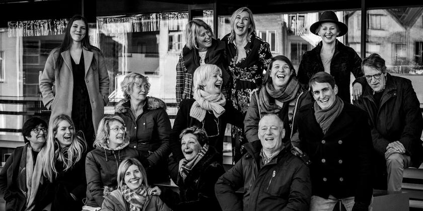 Groepsfoto met 15 lachende mensen, zwart-wit.