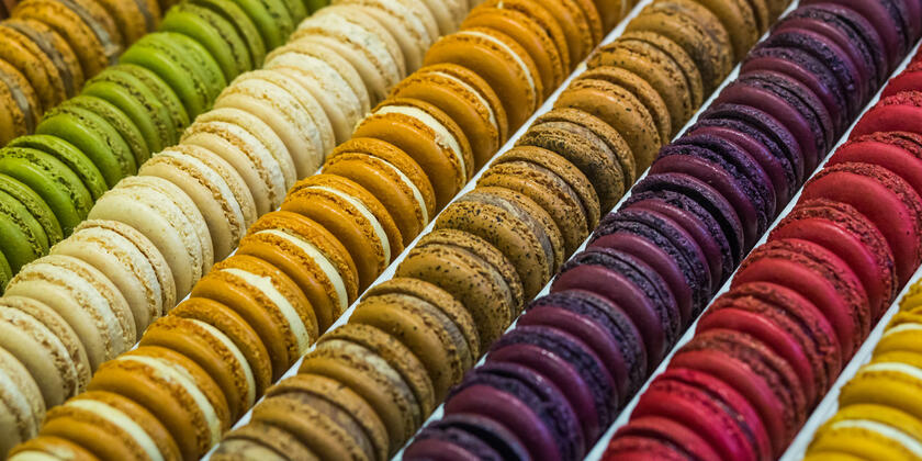 Verschillende rijen gekleurde macarons. Van links naar rechts: Bruin, groen, wit, oker, bruin met zwarte spikkels, paars en rood. 
