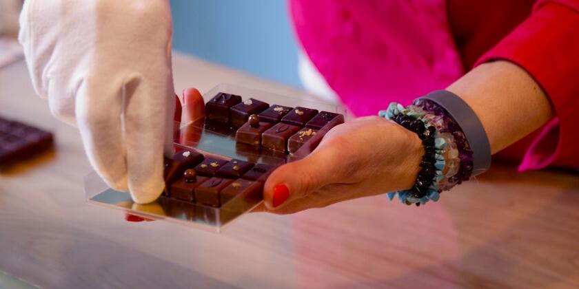 Close-up van een vrouw haar handen die een doosje pralines vast heeft. Aan een hand heeft ze een witte handschoen aan om de chocolade mee vast te nemen.