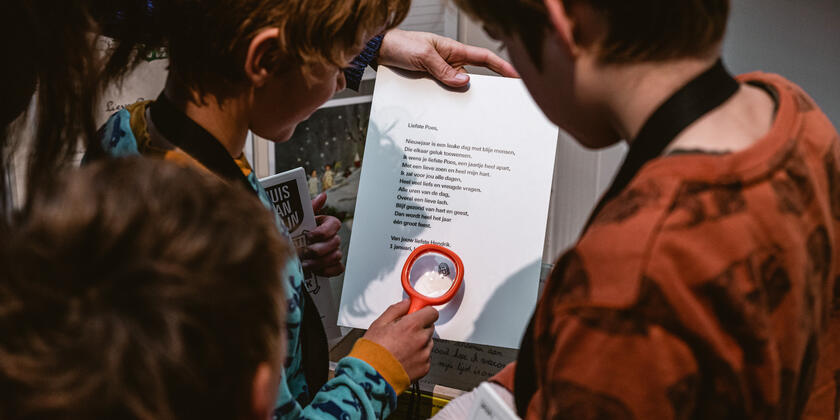 Een groepje kinderen bekijken een nieuwjaarsbrief. Een van hen gebruikt een vergrootglas waardoor je een stempel van een lachend gezichtje kunt zien.