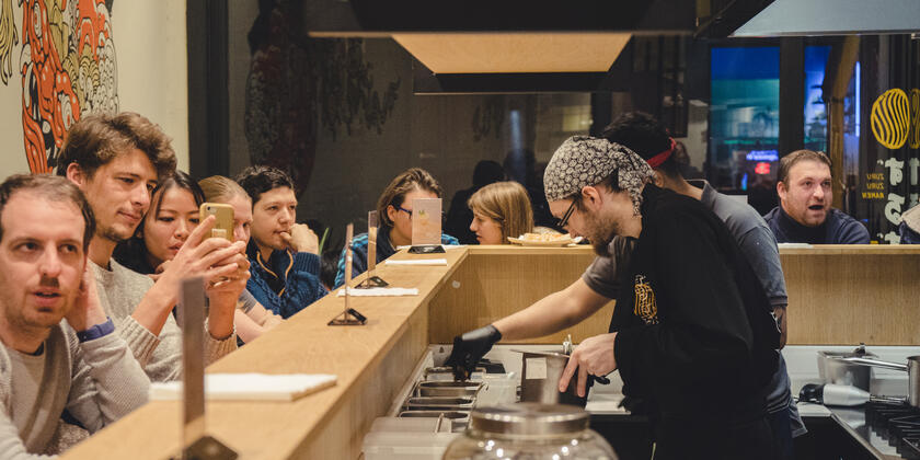 People are sitting at a wooden bar while the chef prepares ramen