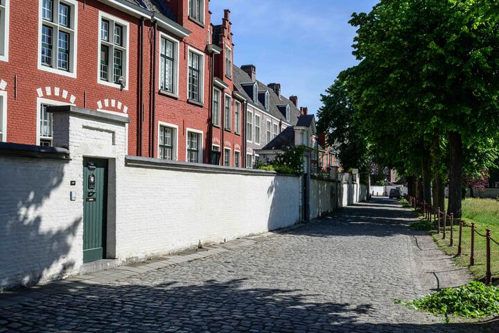 ©VisitGent Kasseipad met aan de linkerkant een witte muur met een groene deur die naar de roodgeverfde huizen van het begijnhof leiden. Aan de rechterkant van het pad staan bomen.
