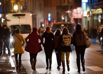Zes studentes stappen weg van de fotograaf richting een drukke straat met cafés en veel mensen, 's nachts.