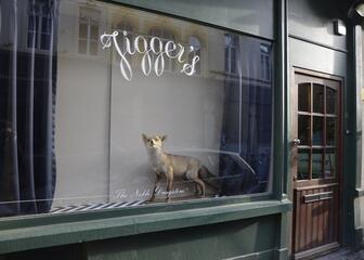Raam met de naam "Jiggers", een opgezette vos in het raam en blauwe gordijnen. Groene gevel en een bruine deur.