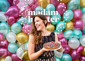 Een bruinharige vrouw die glimlachend een taart in de hand heeft voor een met ballonnen gevulde etalage van Madam Bakster. 