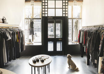 Het interieur van een kledingboetiek. In het algemeen witte muren, maar de deur en etalage zijn zwart geschilderd. Er zit een hond te wachten op klanten. 