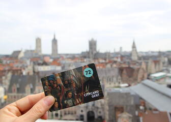 Una mano sujeta la City Card Gent (72h) con una vista panorámica de Gante de fondo