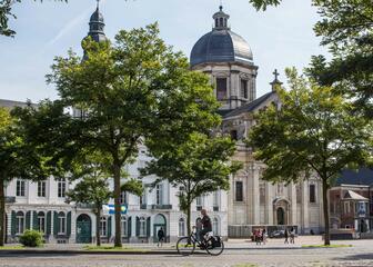 Bild der St.-Peter-Abtei, aufgenommen vom Sint-Pietersplein aus. Im Vordergrund einige Bäume und im Hintergrund die Abtei. Das Bild zeigt auch einen Radfahrer und einige Spaziergänger. 