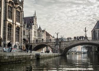Foto van op het water met zicht over de Leie en de Sint-Michielsbrug. Mensen wandelen naast het water en over de brug. Naast het water zie je verschillende middeleeuwse gebouwen.