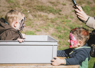 een vrouw die een foto maakt van een geschminkt kind in een doos en een ander geschminkt kind dat de doos vasthoudt