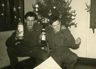 zwart-wit foto van 2 mannen in uniform met elk een fles likeur vast voor een kerstboom