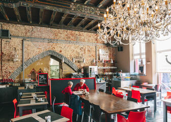 Grote kristallen luchter aan het middeleeuws plafond, bakstenen muren, grote houten tafels en rode stoelen. 