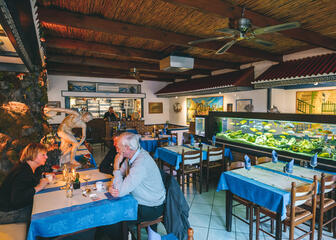 Restaurant met Grieks interieur: houten tafeltjes met blauw tafellaken en witte loper, houten plafonds en een aquarium in het midden van de ruimte.