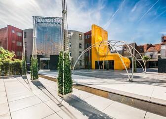 Modern ingericht plein met metalen constructies waar er planten rond groeien, rode, grijze en gele gebouwen op de achtergrond.