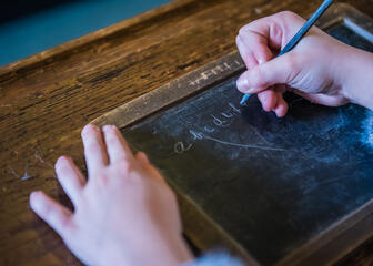 Detail van twee kinderhanden, rechterhand schrijft met griffel op een lei.