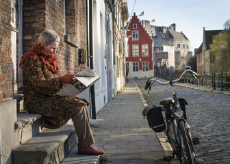 Een vrouw die de krant leest op de dorpel van een woning naast haar fiets.