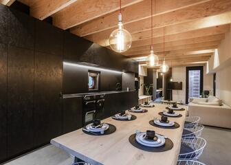 kamer met lange houten en gedekte eettafel, stoelen, zwarte keuken, zithoek met bank en tv