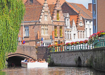 Toeristenbootje vaart richting brug, kades met veel bloemen en trapgeveltjes op de achtergrond.