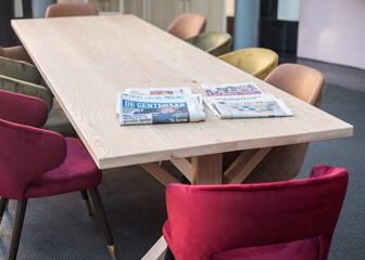 Een grote houten tafel met kleurrijke stoelen (fuchsia, muntgroen, zalmroze, pastelgeel). Op de tafel liggen twee kranten.
