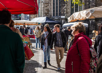 Bezoekers op de brocantemarkt bij Sint-Jacobs. Vrouw met rode jas op de voorgrond, jong koppel op de achtergrond. 