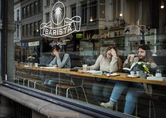 Drie mensen zitten te lezen en koffie te drinken aan de lange tafel bij het raam.