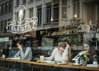 Drie mensen zitten te lezen en koffie te drinken aan de lange tafel bij het raam.